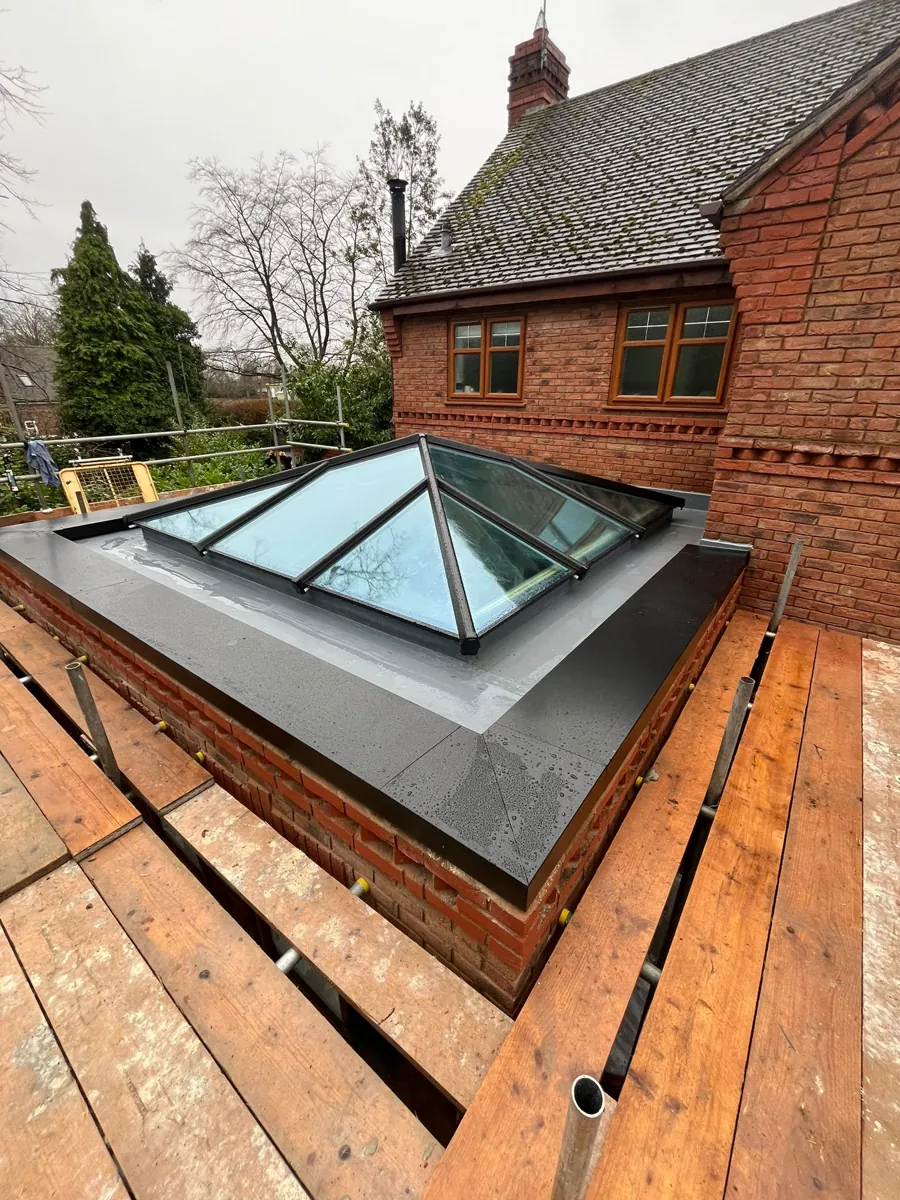 Completed anthracite aluminium roof lantern on a fibreglass flat roof, photographed from the scaffold with the brick house behind